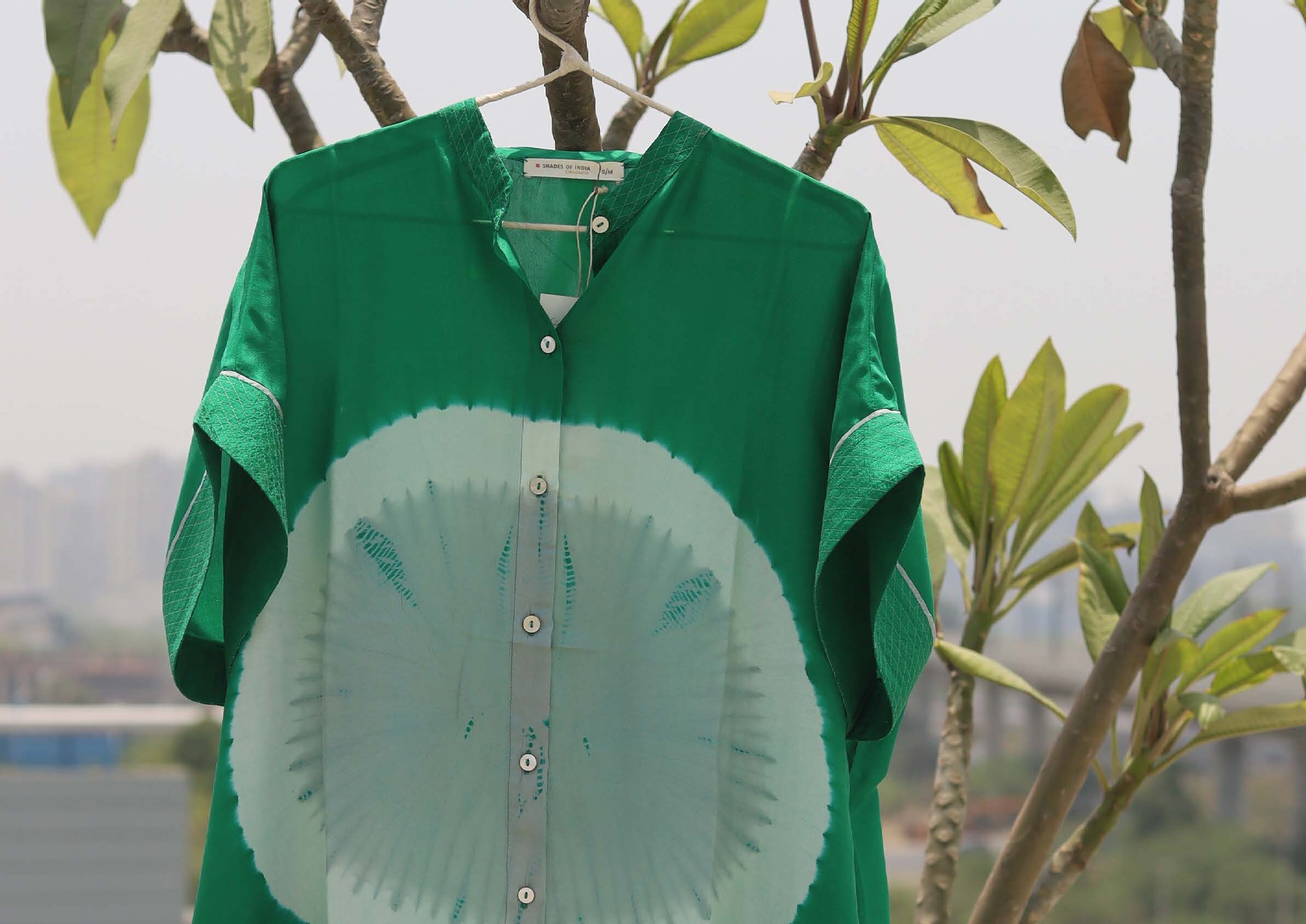 A vibrant green shirt with a unique circular tie-dye pattern is displayed hanging from a branch, highlighting its artistic design against a blurred city backdrop.