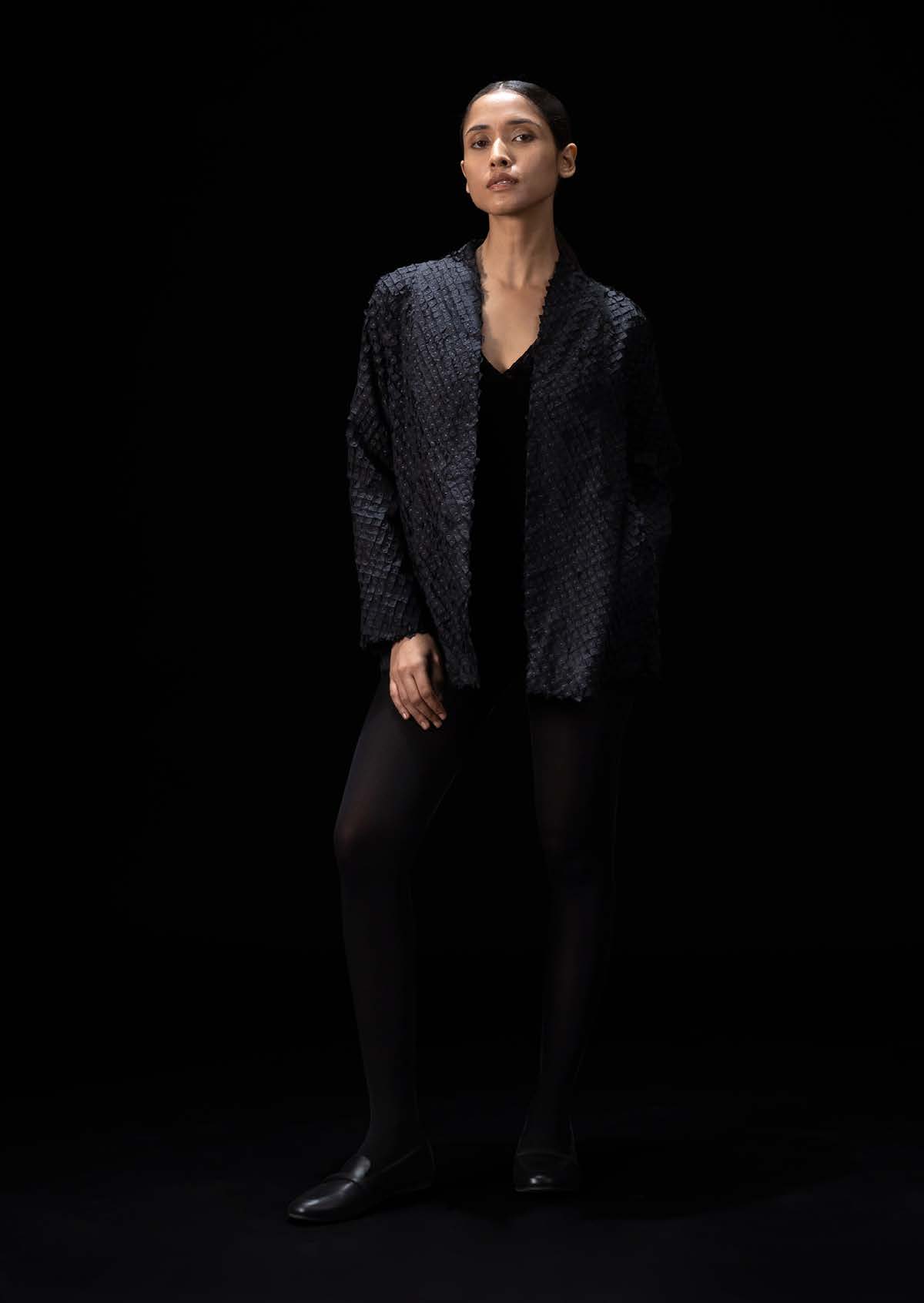 Model in a textured black jacket and leggings poses confidently against a dark background, showcasing minimalist fashion for a contemporary audience.