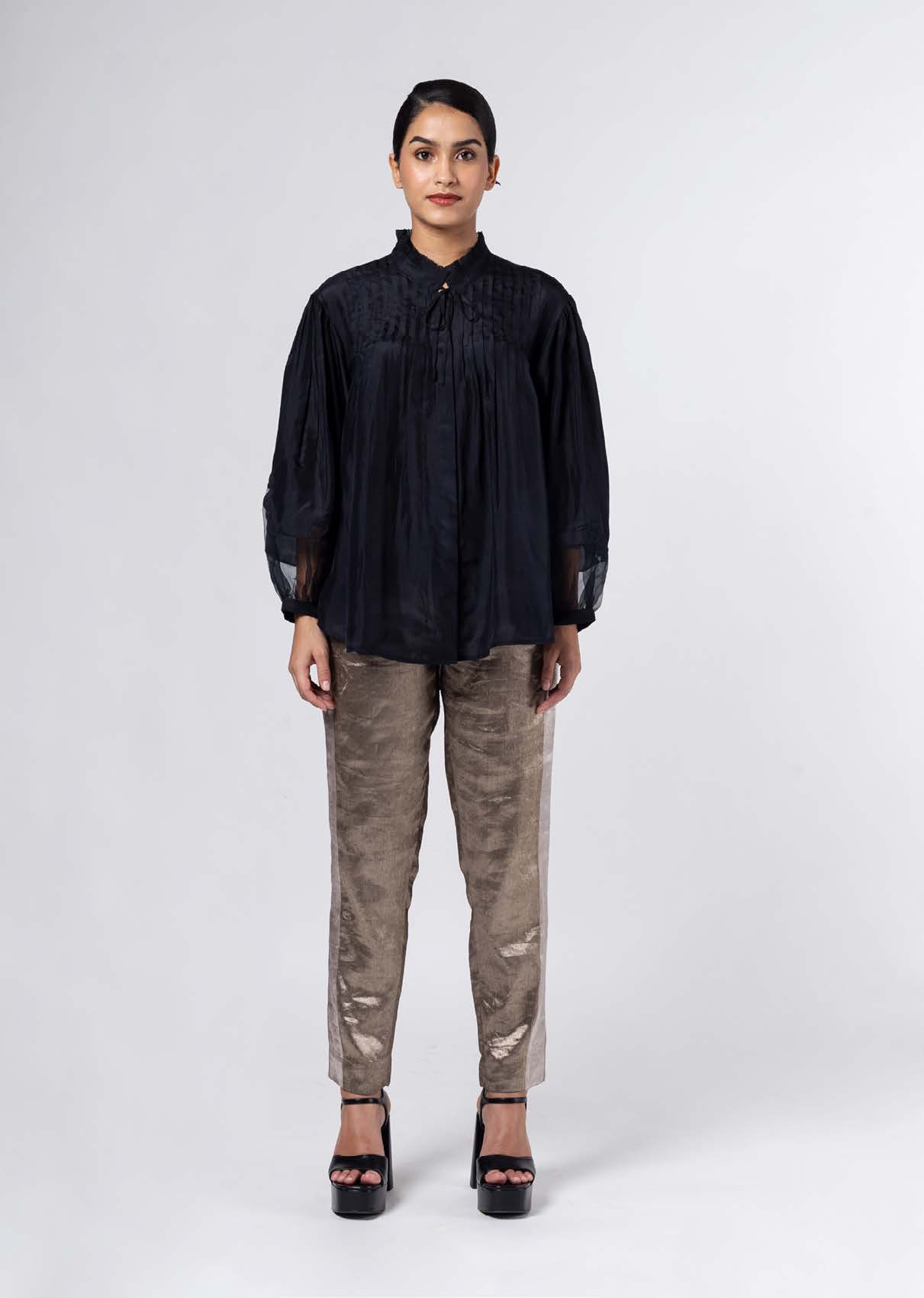 A woman stands confidently in a black pleated blouse and shiny beige pants, complemented by high-heeled black sandals, against a neutral backdrop.