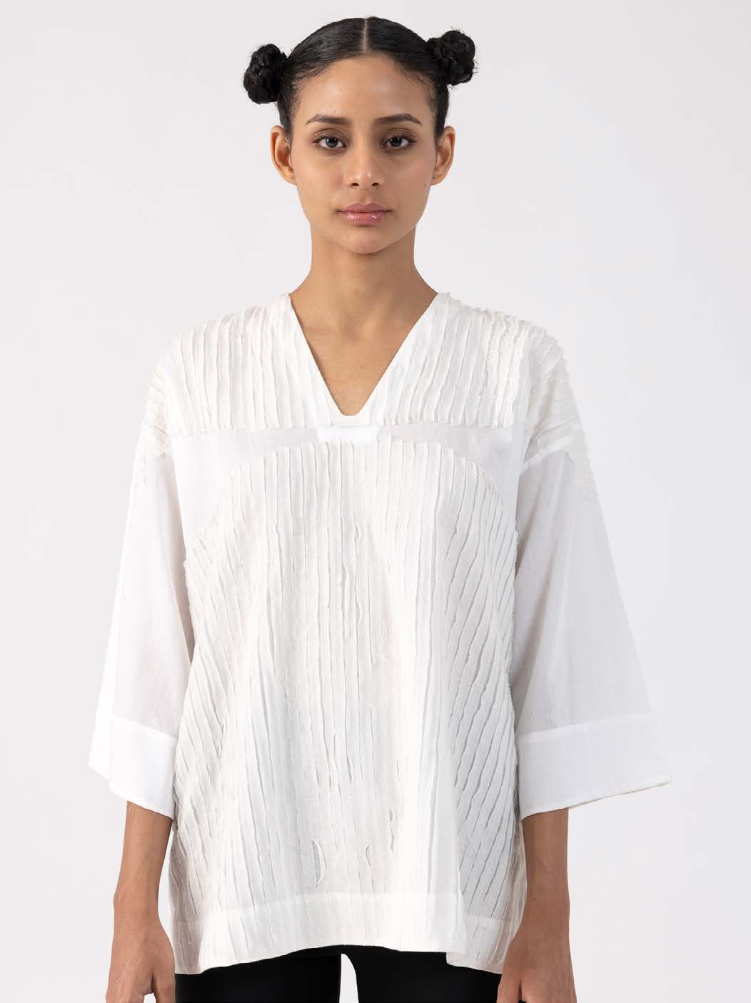 Model wearing a textured white tunic with a V-neckline, featuring loose sleeves and a minimalist design, against a plain background.