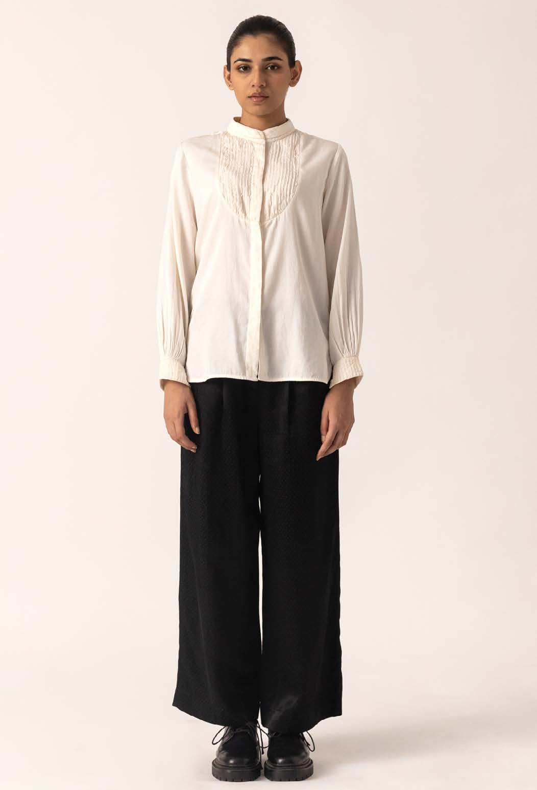 A model stands confidently in a white, button-up shirt with detailing and wide black trousers, showcasing a modern, chic outfit against a neutral background.