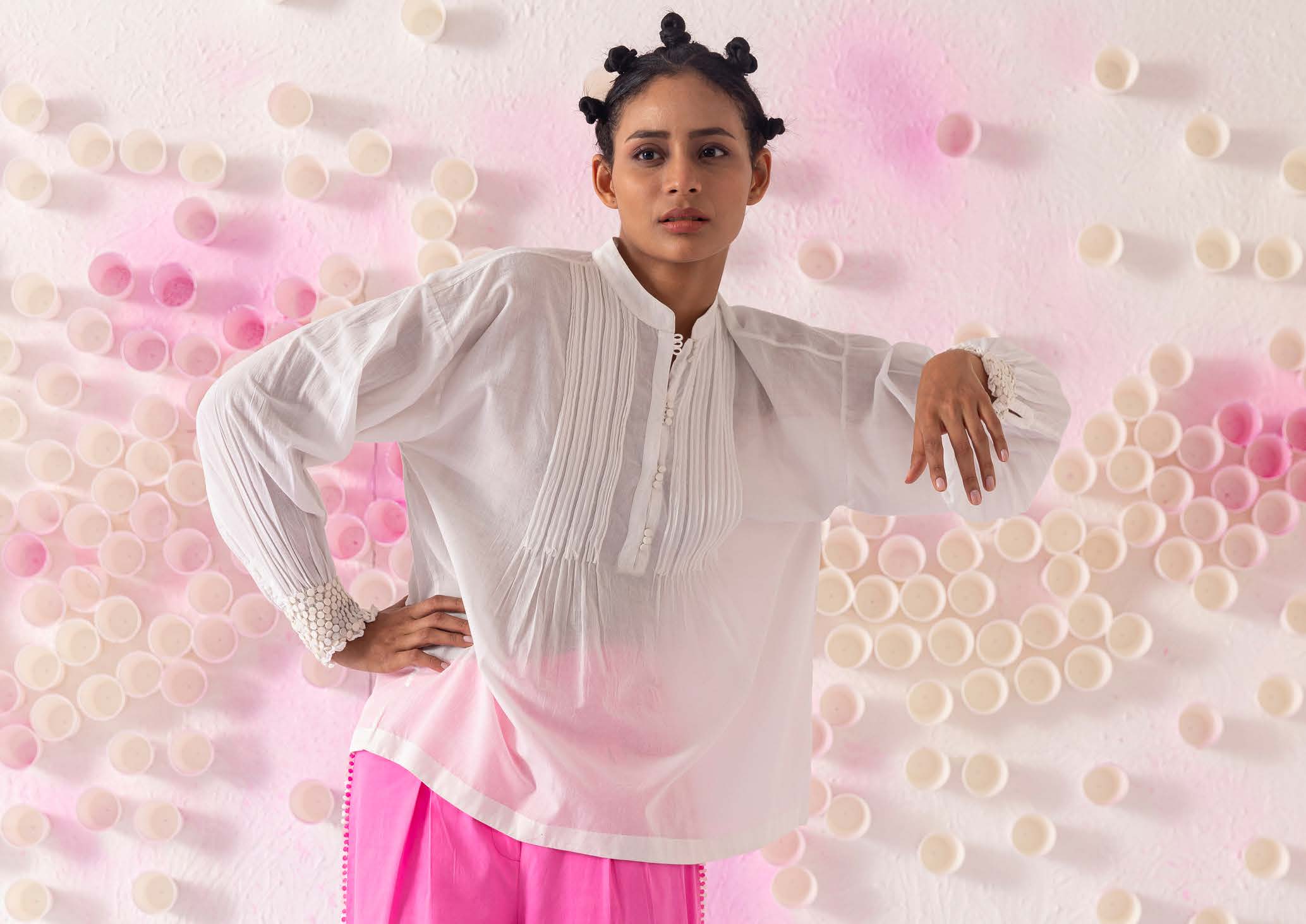 A model poses confidently, wearing a white top and pink skirt, against a backdrop of pastel circles, highlighting contemporary fashion aesthetics.