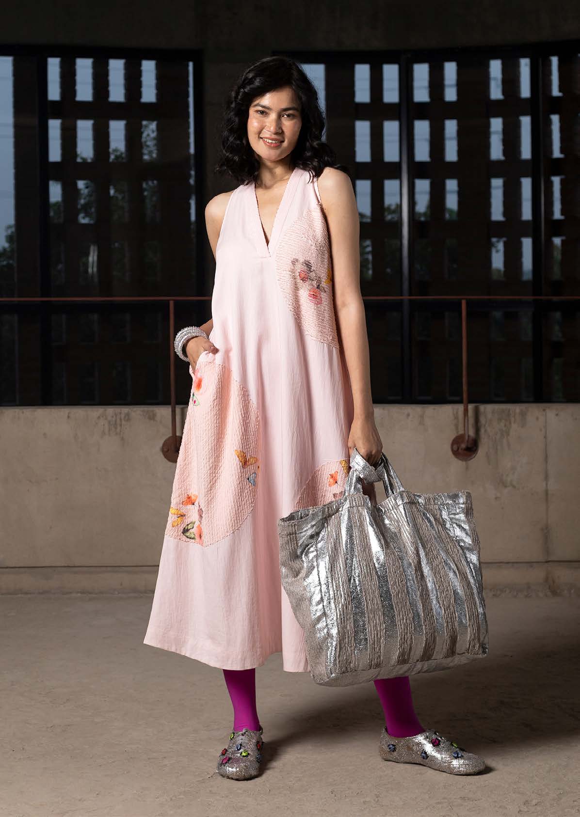 A woman in a pink sleeveless dress, adorned with floral patches, poses confidently, holding a silver tote bag and wearing bright pink tights and playful shoes.