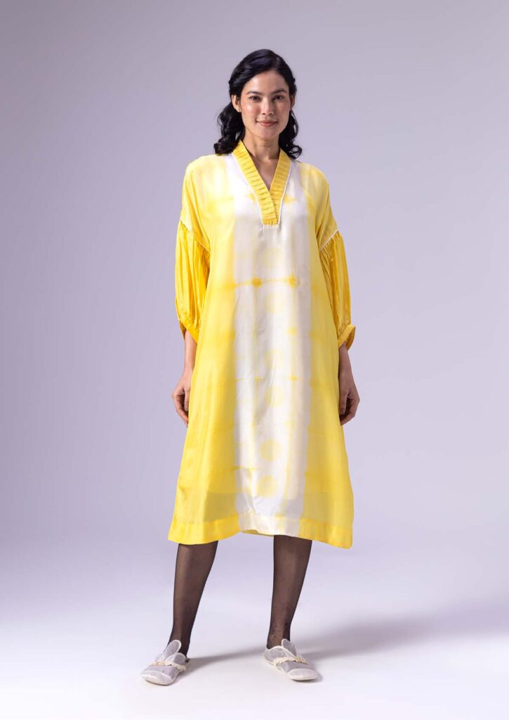 Model wearing a flowing yellow dress with a pleated neckline and balloon sleeves, paired with light slippers, against a neutral background.